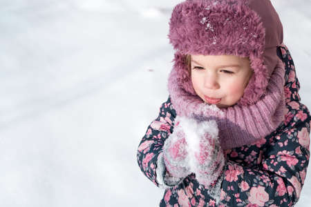 Close-up portrait authentic little preschool minor 3-4 years girl in pink hat warm clothes have fun smiles in snowy frosty weather. Funny kid eat taste snowの写真素材