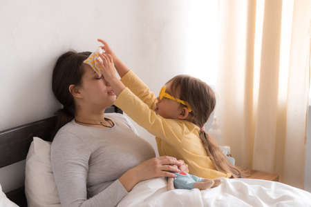 little girl wearing uniform playing doctor, nurse with young mum, nanny in bedroom, checking mother throat, measures temperature, take care, family spend leisure time at home together role play gameの写真素材