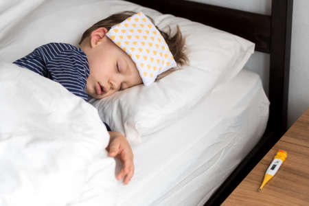 Authentic portrait sick cute caucasian little preschool baby boy in blue sleep with compress on forehead on white bed. child resting at lunchtime. care, medicine and health, Childhood, life conceptの写真素材