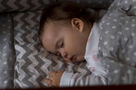 top view Cute little chubby baby girl kid sleeping sweetly in grey crib during lunch rest time in white and pink pajamas with teddy bear at home. Childhood, leisure, comfort, medicine, health conceptの写真素材