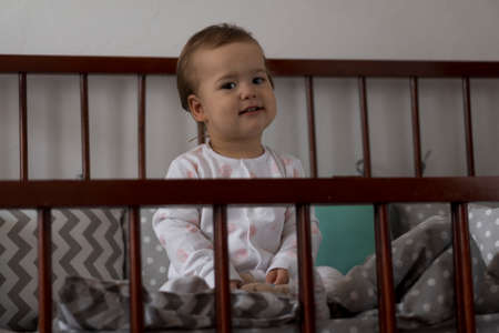 Cute cheerful little chubby baby girl sitting in grey baby crib after waking up from sleep in white and pink pajamas with teddy bear and smiling. Childhood, leisure, comfort, medicine, health conceptの写真素材