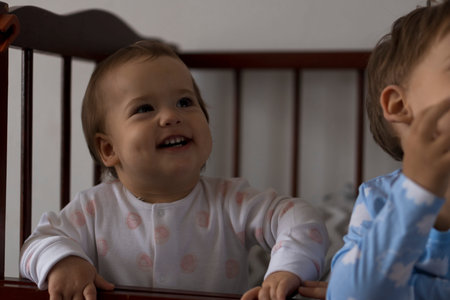 Cute cheerful little chubby baby girl sitting in grey baby crib after waking up from sleep in white...