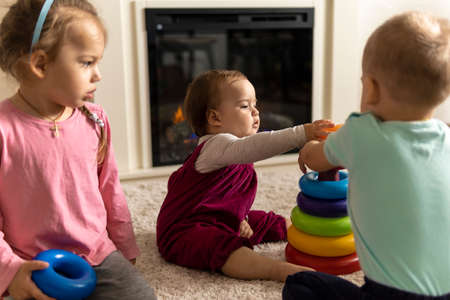 family, friends, friendship, childhood, development, skills, motility games - little authentic children toddler kids puts rings on pyramid playing together with toy sit on floor indoors at cozy homeの写真素材