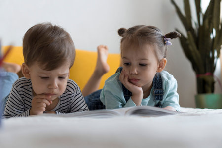 Two preshool, toddler children read large interesting book of fairy tales on yellow bed. Siblings...