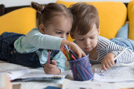 smart smiling preshool toddler children boy and girl draw with pencils lying on their stomach at yellow bed. little cute reader have fun, happy kid on quarantine at home. Friendship, family, educationの写真素材