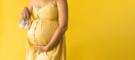 Motherhood, femininity, fashion, hot summer - banner Close-up unrecognizable pregnant woman lady in beautiful vintage dress Provence straw hat with small baby shoes on yellow background, copy spaceの写真素材