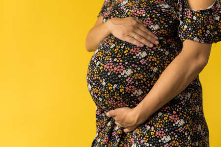 Motherhood, femininity, love, care, waiting, hot summer - bright croped Close-up unrecognizable pregnant woman in floral black dress with hands over tummy rub belly on yellow background, copy spaceの写真素材