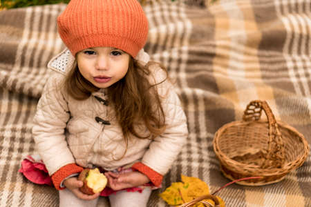 Close Up Portrait Of Little Cute Preschool Minor Girl In Orange Beret On Yellow Fallen Leaves Eats Red Apple Look At Camera Cold Weather In Fall Park. Childhood, Family, Motherhood, Autumn Conceptの写真素材