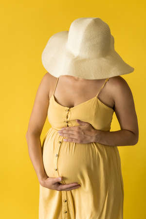 Motherhood, femininity, fashion,, hot summer - croped Close-up unrecognizable pregnant woman lady in beautiful vintage dress Provence straw hat with hands over tummy rubs tummy on yellow backgroundの写真素材