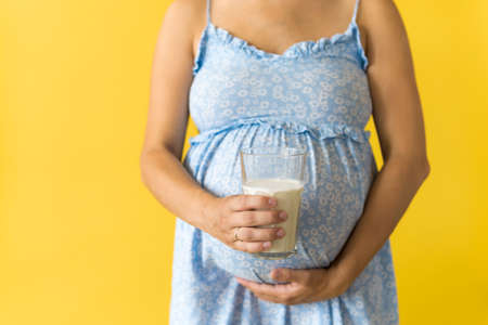 Motherhood, femininity, vitamins, calcium, healthy eating food, dairy, hot summer - croped portrait pregnant unrecognizable woman in floral blue dress holds drink glass of milk on yellow backgroundの写真素材