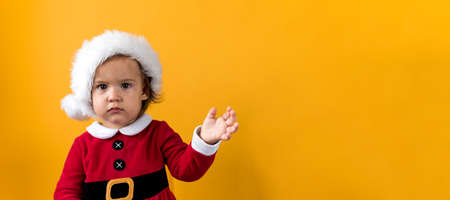 Banner Serious Portraite Cute Fun Cheerful Chubby Baby Girl in Santa Hat Waving Hand At Yellow Background. Child Play Christmas Scene Celebrating Birthday. Kid Have Fun Spend New Year Time Copy Spaceの写真素材