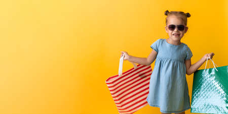Banner Portrait Beautiful Happy Little Preschool Girl In Sunglasses Smiling Cheerful Holding Cardboard Bags Isolated On Orange Yellow Background. Happiness, Consumerism, Sale People shopping Conceptの写真素材