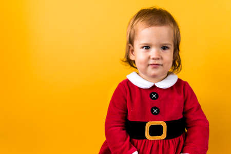 Banner Portraite Cute Happy Cheerful Chubby Baby Girl in Santa Suit Looking On Camera At Yellow Background. Child Play Christmas Scene Celebrating Birthday. Kid Have Fun Spend New Year Time Copy Spaceの写真素材