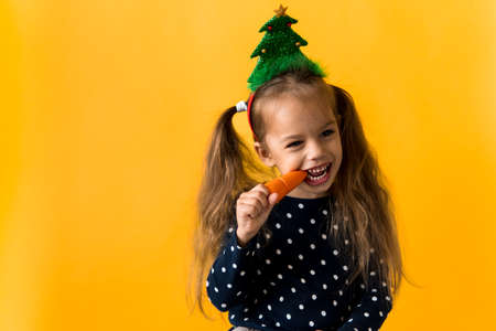 Portrait positive cheerful smiling happy little schoolgirl girl Christmas tree decoration polka dot dress biting eat orange carrots on orange background. New year, holiday, celebration, winter conceptの写真素材