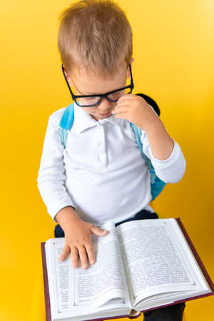 Portrait of Funny Preschool Child Boy in Big Glasses sits on chair Reads Book on Yellow Background. Happy Smiling Kid Go Back to School, Kindergarten. Success, Motivation, Genius, Superhero conceptの写真素材