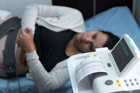 Pregnant Woman On Hospital Couch During Medical Control Cardiotocography. Gynecologist Doctor Hands Turn On Adjust CTG Device Clinic Consultant. Examine Expectant Belly Baby Mother Healthcare Check Upの写真素材
