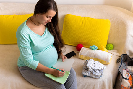 Cute Pregnant Woman writing packing list for maternity hospital With Notebook Prepares bags. Young...