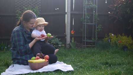Happy Young Cheerful Mother Holding Baby Eating Fruits On Green Grass. Mom Adorable Infant Child...
