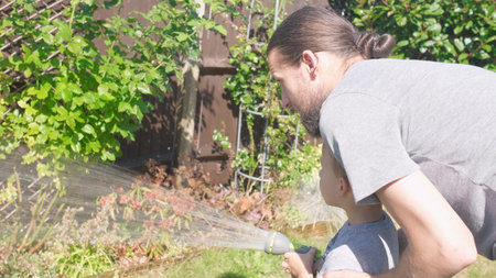 Father and Son. Funny little boy watering lawn plants in garden housing backyard with Dad. Summer...