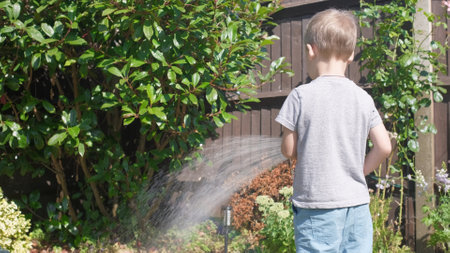 Funny little boy watering lawn plants in garden housing backyard. Adorable child playing with irrigation hose at hot sunny summer outdoors. Children help with housework. activity for kids. Childhoodの写真素材
