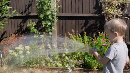 Funny little boy watering lawn plants in garden housing backyard. Adorable child playing with irrigation hose at hot sunny summer outdoors. Children help with housework. activity for kids. Childhoodの写真素材