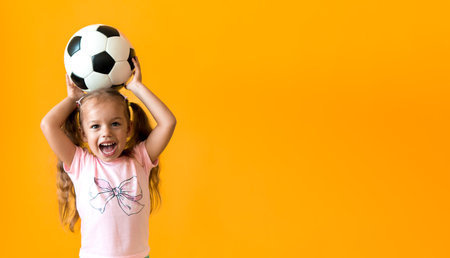 Authentic cute smiling preschool little girl with classic black and white soccer ball look at camera on yellow background. child play football in t-shirt and shorts. Sport, championship, team conceptの写真素材