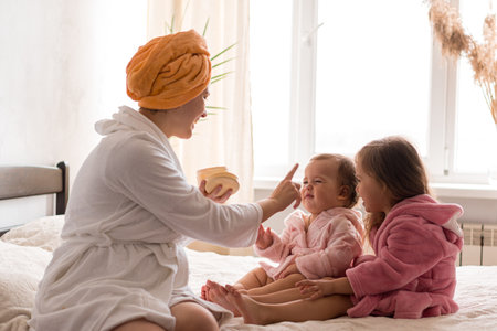 Young mother two daughters in curlers bathrobes. happy smiling family skin care concept. Mom teaches little kids child to do makeup smear cream on face sitting in orange bathrobes on bed in bedroomの写真素材