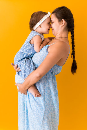 Motherhood, hot summer - portrait young pregnant happy smiling joy mother woman in blue dress holding little baby daughter toddler sibling bare feet in arms looking into eyes kiss on yellow backgroundの写真素材