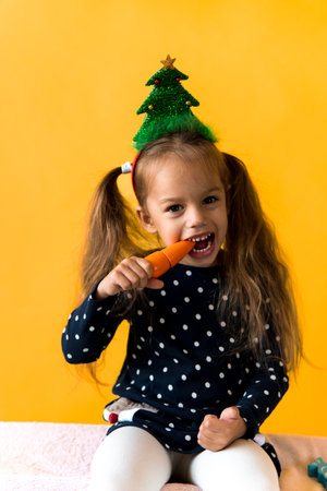 Portrait positive cheerful smiling happy little schoolgirl girl Christmas tree decoration polka dot dress biting eat orange carrots on orange background. New year, holiday, celebration, winter conceptの写真素材