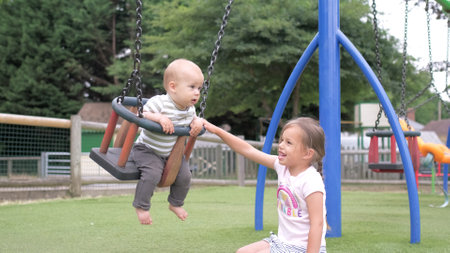 Funny Little Preschool Chubby Girl Older Sister Rides Younger Newborn Infant Brother Boy On Swing. Baby Playing On Playground. Kids Entertainment, Childhood, Child Development, Happy Family Conceptの写真素材
