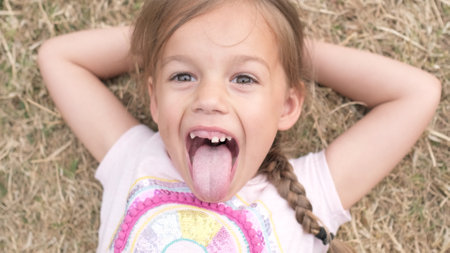 Little Happy Toothless Smiling Child Girl Laying on Yellow Lawn Dry Grass Hay in Park Shows Tongue. Summer Time, Nature, Lifestyle Country life farm village. Portrait Face Close up Looking at Cameraの写真素材