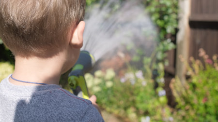 Funny little boy watering lawn plants in garden housing backyard. Adorable child playing with irrigation hose at hot sunny summer outdoors. Children help with housework. activity for kids. Childhoodの写真素材