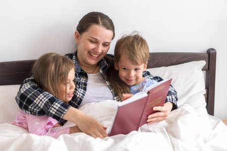 happy family. Close up loving mother lying with daughter son two kids pajamas in bed children reading interesting storybook, preschool smiling kid girl enjoy fairytale before go to sleep good pastimeの写真素材