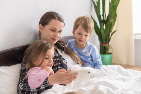Happy Young Woman Mother With 3-4 Aged Children Siblings Baby Boy And Girl Watching Smartphone Together Digital Tablet On White Bed. Mom With Daughter And Son Family Making Selfie In Pajamas.の写真素材