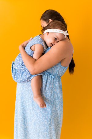 Motherhood, hot summer - portrait young pregnant happy smiling joy mother woman in blue dress holding little baby daughter toddler sibling bare feet in arms looking into eyes kiss on yellow backgroundの写真素材