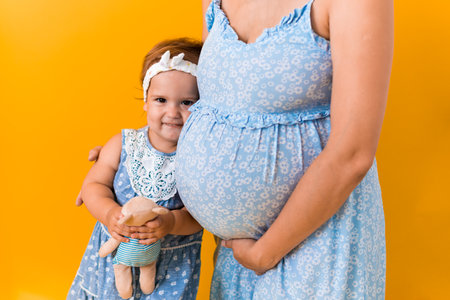 Motherhood, femininity, hot summer - croped portrait pregnant unrecognizable woman mother in blue dress little happy girl baby sibling with teddy bear hugs care moms big belly on yellow backgroundの写真素材