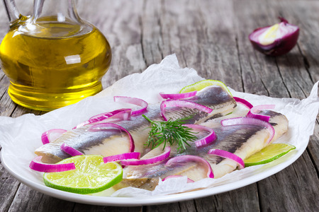 herring fillets with onion and lime slices on parchment paper on an oval dish on an old rustic table with a bottle of olive oil , close-upの写真素材