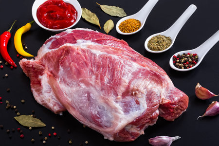 fresh raw pork shoulder with ingredients for marinade: Black, green, pink and white peppercorns, dried herbs, chili, bay leaf, garlic, barbecue sauce on black background, view from above, close-upの写真素材