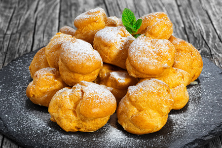 Delicious profiteroles filled with cream on slate plate,  dusting of powdered sugar and decorated with mint, on old dark wooden table, close-upの写真素材
