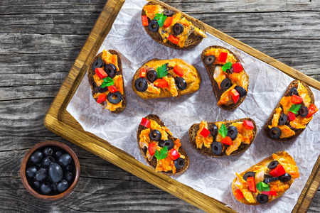 Crostini with pieces of fish stewed in tomato sauce, rings of black olives, red bell pepper, on white paper on chopping board, view from aboveの写真素材