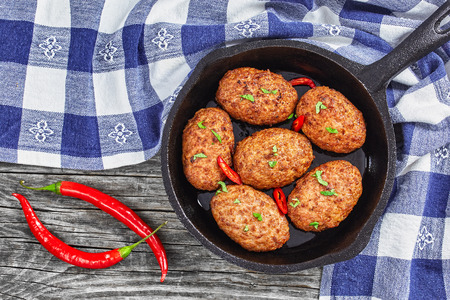 juicy delicious meat cutlets in iron frying pan sprinkled with chili pieces and green herbs. tomato, chili peppers on old rustic planks, view from above, close-upの写真素材