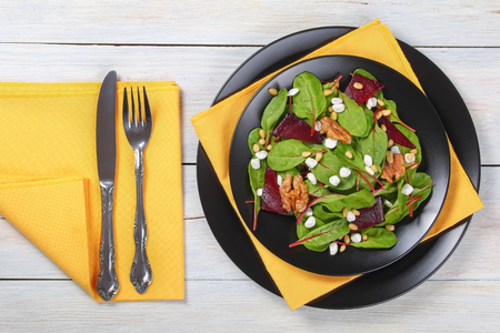 fresh delicious healthy salad of chard leaves, granulated cottage cheese, baked beetroot slices and nuts on black platters, on white wooden planks, with cutlery on napkin,  view from aboveの写真素材