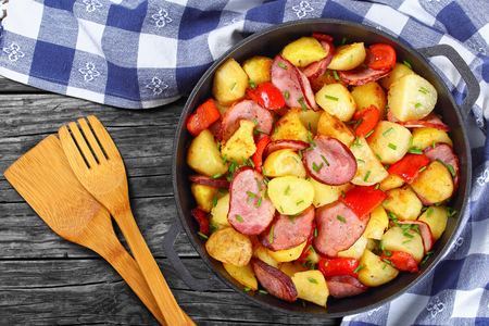 tasty roasted potato wedges with ham and red bell pepper, sprinkled with fresh green spring onion in iron pan on dark rustic table with kitchen towel, view from above, german styleの写真素材