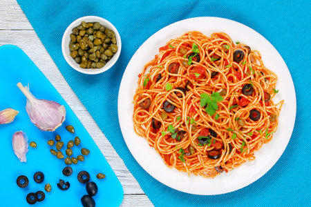 Pasta spaghetti with tomato sauce, capers and olives on plate with ingredients on cutting board on  wooden background, traditional Italian recipe view from aboveの写真素材