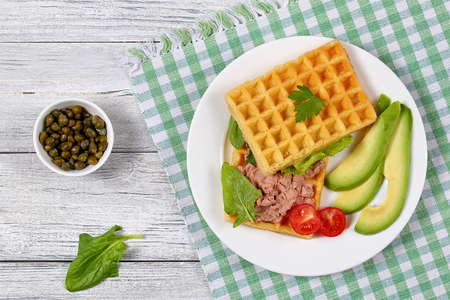 delicious salted waffles sandwich with avocado slices, turkey liver, meat pate, lettuce, spinach, cherry tomatoes and parsley on white plate, capers in bowl on background, view from aboveの写真素材