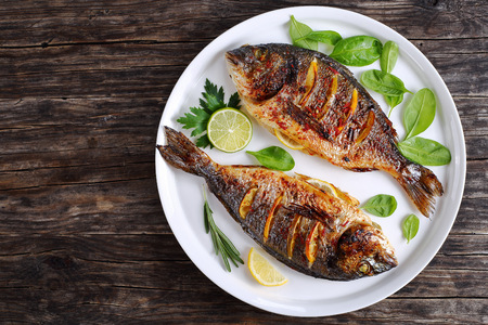 delicious roasted dorado or sea bream fish with lemon and orange slices, spices, fresh parsley and spinach on white platter on old dark wooden table view from aboveの写真素材