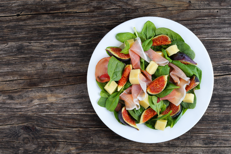 spinach, figs, thinly sliced italian ham and tender mozzarella cheese salad on white plate on dark wooden kitchen table, view from aboveの写真素材