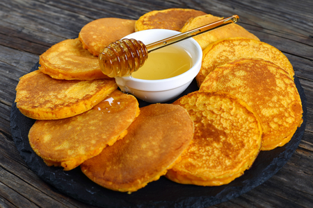Delicious golden yellow pumpkin pancakes on black stone plate with honey in center on dark wooden table, view from above, close-upの写真素材