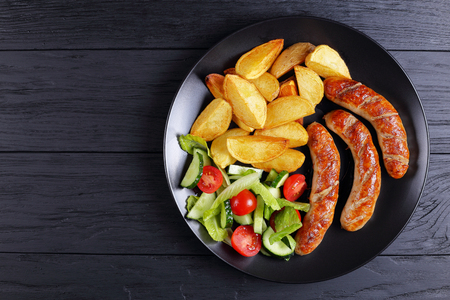 delicious hot grilled sausages, baked potatoes and fresh vegetables romaine lettuce salad on black plate on black wooden boards, view from aboveの写真素材