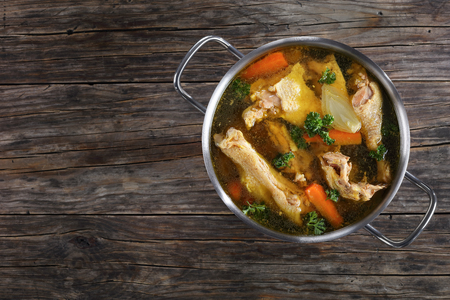 clear Chicken broth with pieces of rooster meat on bone and vegetables in a metal casserole on dark wooden table, view from aboveの写真素材